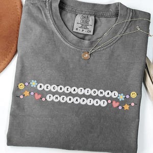 Op de afbeelding: Een grijs Comfort Colors T-shirt met de witte woorden "RECREATIONAL THERAPIST", versierd met smiley's, bloemen, sterren en harten. Een gouden ketting en een bruine hoed zijn ook zichtbaar.