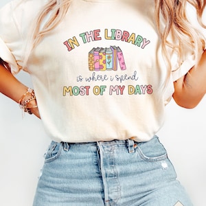 May include: A white t-shirt with the text "In the Library is where I spend most of my days" in rainbow colours. The text is surrounded by a stack of colourful books with flowers and hearts.