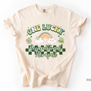May include: A cream colored t-shirt with a green, yellow, and pink rainbow, shamrocks, and a leprechaun hat. The text "One Lucky Occupational Therapist" is printed on the shirt.