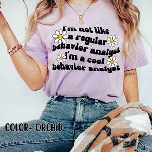 K&ouml;nnte beinhalten: Ein lila T-Shirt mit einem G&auml;nsebl&uuml;mchen-Motiv und dem Text "I'm not like a regular behavior analyst I'm a cool behavior analyst". Der Text ist in einem Retro-Schriftart-Stil. Das Model tr&auml;gt eine blaue Jeans und einen karierten Schal.