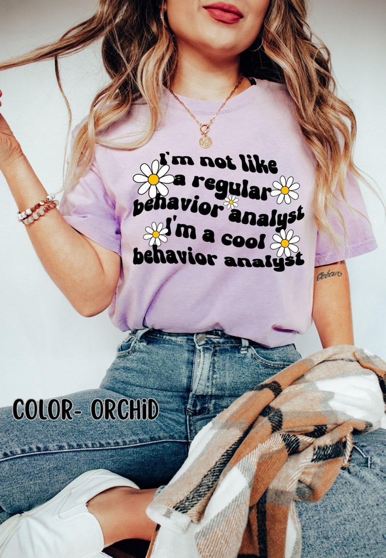 K&ouml;nnte beinhalten: Ein lila T-Shirt mit einem G&auml;nsebl&uuml;mchen-Motiv und dem Text "I'm not like a regular behavior analyst I'm a cool behavior analyst". Der Text ist in einem Retro-Schriftart-Stil. Das Model tr&auml;gt eine blaue Jeans und einen karierten Schal.