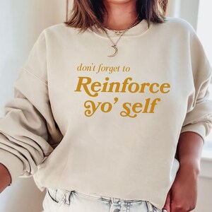 May include: A cream-colored sweatshirt with the text "don't forget to Reinforce yo' self" in a gold-colored font.