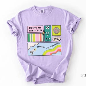 May include: Orchid-colored t-shirt with a comic strip design. The design includes the text "DOING MY BEST CLUB", cartoon faces, a globe, and the phrase "PROGRESS NOT PERFECTION". The shirt is made of a soft material.