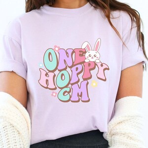 Puede incluir: Una camiseta de color lila claro con las palabras "ONE HOPPY CM" en una fuente retro, con un conejito de dibujos animados y flores. La camiseta es de color pastel y tiene mangas cortas.