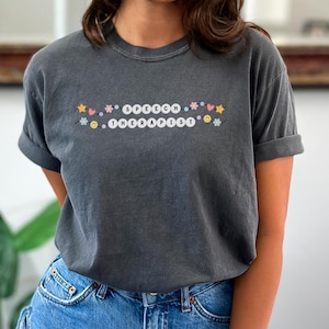 Könnte beinhalten: Ein dunkelgraues T-Shirt mit dem Text "SPEECH THERAPIST" in weißen Buchstaben, umgeben von bunten Cartoon-Symbolen.
