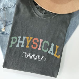 May include: A gray t-shirt with the text "PHYSICAL THERAPY" in colorful letters. The letters are in a rainbow color scheme with a white outline.