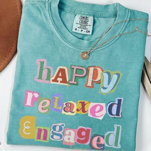 Op de afbeelding: Zeegroen t-shirt met de woorden "Happy, Relaxed, Engaged" in een kleurrijke, uitgesneden stijl. Een gouden ketting met een hanger ligt op het shirt. Een bruine hoed en een gedroogde plant staan op de achtergrond.