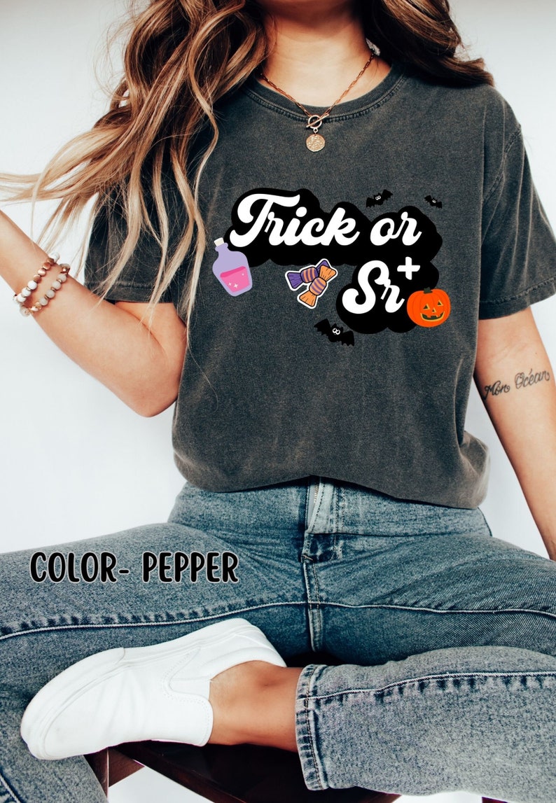 Puede incluir: Una camiseta gris oscuro con el texto "Trick or Sr+" en blanco y negro. La camiseta tiene un gr&aacute;fico de una botella de poci&oacute;n, caramelos, un murci&eacute;lago y una calabaza.