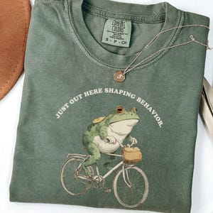 May include: Moss green t-shirt featuring a frog on a bicycle graphic. The frog is green and white, with orange eyes and a tan bag on the handlebars. The text "JUST OUT HERE SHAPING BEHAVIOR" is arched above the frog. A gold necklace is draped across the shirt.