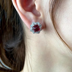 May include: A close-up of a stud earring with a square, deep red gemstone at its centre, encircled by a halo of smaller, clear gemstones. The earring is set against a person's ear and hair.