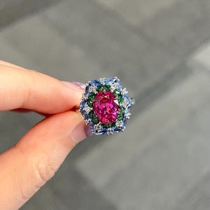 May include: A silver-toned ring featuring a central oval-cut vibrant pink gemstone, surrounded by a halo of marquise-cut green stones, an outer border of light blue marquise-cut stones, and small clear accent stones set in a metallic frame.