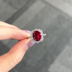 May include: A ruby-colored gemstone ring with a halo of small, clear stones. The ring features a large, oval-cut ruby at the center, surrounded by a cluster of smaller, round-cut diamonds. The band is thin and silver-colored.
