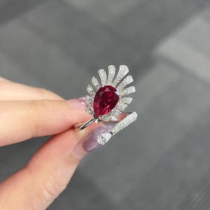 Könnte beinhalten: Ein silberfarbener offener Ring mit einem großen, birnenförmigen, tiefroten Edelstein in der Mitte, umgeben von einer fächerförmigen Anordnung kleiner, klarer Pavé-gefasster Steine. Ein einzelner, kleinerer birnenförmiger Stein ziert das andere Ende.