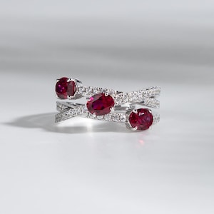 Peut inclure: Une bague en argent ornée de trois rubis ovales et de nombreux petits pierres précieuses transparentes. Le design de la bague présente des bandes entrecroisées, créant un effet superposé. Les rubis sont d'un rouge profond.