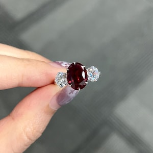 May include: A three-stone ring featuring a large, oval-cut deep red gemstone at the center, flanked by two smaller, round-cut clear white gemstones. The stones are set in a polished silver-toned metal band with visible prongs securing each gem.