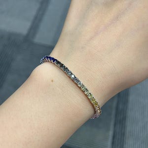 May include: A tennis bracelet featuring a continuous row of small, round-cut gemstones arranged in a gradient colour pattern. The stones transition in hue from deep blue and dark grey to light grey, pale yellow, and vibrant yellow along the metal band.