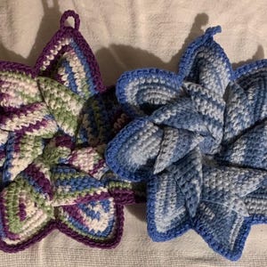 May include: Two star-shaped crocheted potholders with hanging loops. One potholder features a variegated yarn pattern in shades of purple, green, and white. The second potholder displays a variegated yarn pattern in various shades of blue and white.