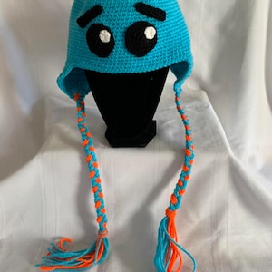 May include: A handmade, blue crochet hat with a cartoon face design. The hat features large black eyes with white highlights, black eyebrows, and orange yarn accents. Braided ear flaps with orange and blue tassels complete the whimsical look.