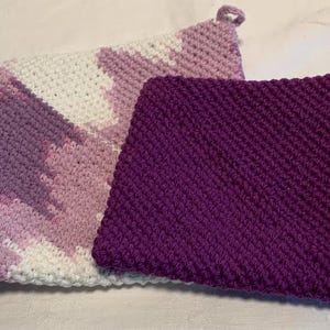 May include: Two square crochet potholders with hanging loops. One potholder features a variegated pattern of white, light purple, and dark purple yarn. The second potholder is a solid, deep purple color with a textured, dense stitch pattern throughout.