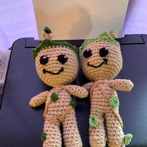 May include: Two crocheted plush toys of the character Groot from the Guardians of the Galaxy franchise. The toys are beige with green leaves and black eyes. They are smiling and have a happy expression.