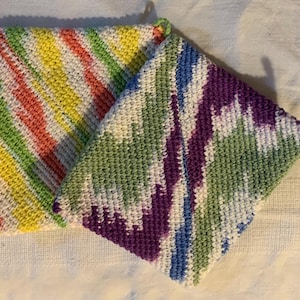 May include: Two square crocheted potholders featuring textured yarn patterns. One potholder displays diagonal stripes of yellow, green, and coral on a white base. The second potholder features a variegated pattern of purple, blue, and green on white.