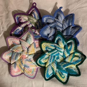 May include: Four star-shaped crochet potholders arranged in a cluster. Each potholder features a unique color palette, including shades of purple, blue, pastel pink, yellow, and teal, all crafted with a textured, multi-colored yarn pattern.