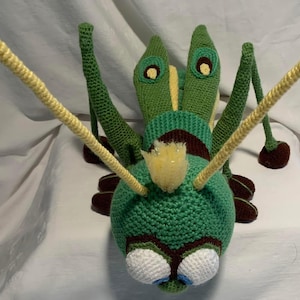 May include: A handmade crochet insect toy in shades of green, yellow, brown, white, and blue. The toy features large eyes, long antennae, and multiple legs. The insect is lying on its back, showcasing its detailed crochet work.