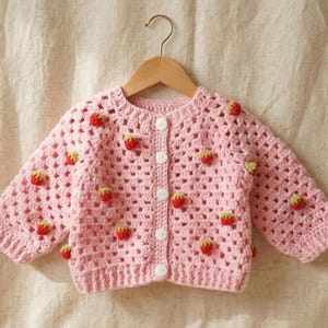 Peut inclure: Un cardigan rose clair pour bébé, crocheté et orné d'applications de fraises rouges et jaunes. Le pull à boutons est présenté sur un cintre en bois, sur fond neutre. C'est un article fait à la main.