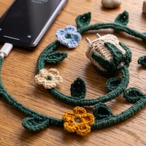 May include: A crocheted charger cable with a floral design. The dark green cable is adorned with leaves and colorful flowers in blue, orange, and beige. The charger is plugged into a black smartphone on a wooden surface.