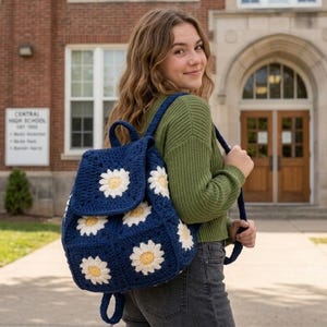 Può includere: Uno zaino all'uncinetto blu navy con disegni di fiori di margherita bianchi e gialli. Lo zaino ha spallacci regolabili e una chiusura a patta. La persona indossa un maglione verde e jeans scuri. Lo sfondo è un edificio in mattoni.