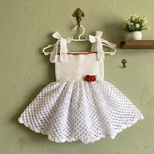 Könnte beinhalten: Ein weißes Häkelkleid mit einem Oberteil mit geradem Ausschnitt, zwei zum Binden geeigneten Schulterträgern und einem ausgestellten Rock mit Lochmuster. Ein kleines Bündel roter Stoffblumen ist an der rechten Seite des Oberteils befestigt.