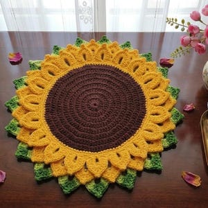 Può includere: Un centrino a forma di girasole all'uncinetto in giallo, marrone e verde, appoggiato su un tavolo di legno scuro. Petali di rosa sparsi circondano il centrino. Un vaso di fiori rosa e un piccolo vassoio con attrezzi sono anche sul tavolo.