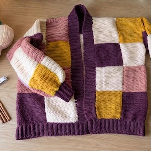 Pode incluir: Um cardigan patchwork feito &agrave; m&atilde;o em tons de roxo, branco, rosa e amarelo. O su&eacute;ter aberto &eacute; exibido com novelos de l&atilde;, agulhas de croch&ecirc;, um caderno, uma caneta, uma fita m&eacute;trica e tesouras. O cardigan &eacute; uma pe&ccedil;a de roupa aconchegante e artesanal.