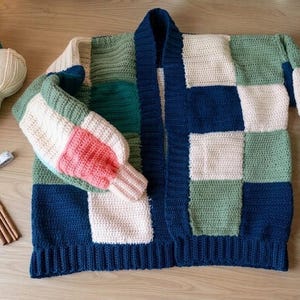 Pode incluir: Um cardigan de croch&ecirc; patchwork feito &agrave; m&atilde;o em tons de azul, verde e creme. O su&eacute;ter de frente aberta apresenta bainha e punhos canelados, com uma mistura de pain&eacute;is quadrados e retangulares. Fios, agulhas de croch&ecirc; e um caderno est&atilde;o por perto.