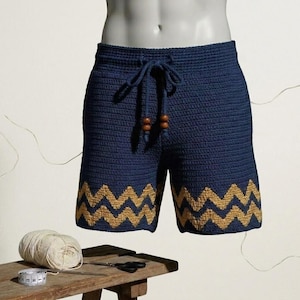 May include: Navy blue crochet shorts with a drawstring waist and a gold zigzag pattern along the hem. The shorts have a relaxed fit and are made of a textured yarn. The drawstring has wooden beads.