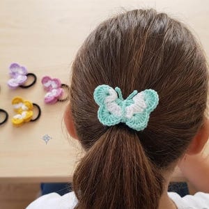 May include: A collection of crochet butterfly hair ties in various colours including light blue, yellow, purple, and pink. Each butterfly is attached to a black elastic hair band. One light blue and white butterfly hair tie is worn in hair.