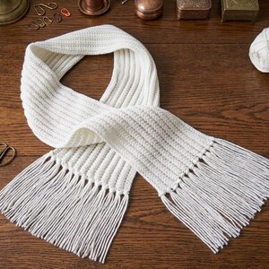 May include: A cream-colored knitted scarf with fringe, lying on a wooden surface. The scarf is next to several balls of white yarn, a crochet hook, and small scissors. The scene suggests a handmade craft project.