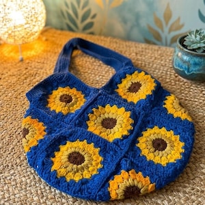 May include: A blue crochet tote bag featuring a sunflower design. The bag has a rectangular shape with a handle and is decorated with yellow and brown sunflowers. The bag is sitting on a woven surface.