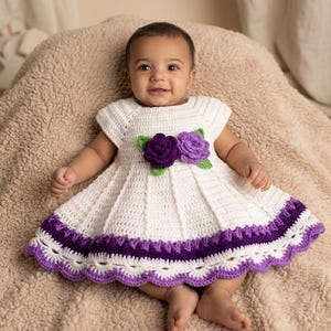 May include: A baby wearing a white crochet dress with purple and green floral accents. The dress features a pleated skirt and a scalloped hem. The baby is lying on a soft, beige blanket.