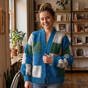 Pode incluir: Uma mulher usando um cardigan patchwork em tons de azul, verde e branco. O su&eacute;ter aberto tem decote em V e mangas compridas. O fundo inclui uma estante e uma janela com plantas.