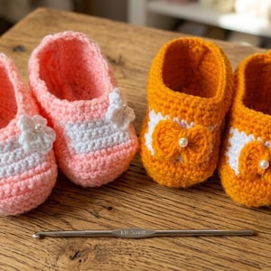 Könnte beinhalten: Zwei Paar handgefertigte Babyschuhe. Ein Paar ist rosa mit weißen Akzenten und einer Blumenverzierung. Das andere Paar ist orange mit weißen Akzenten und einer Schleife mit einer Perle. Ein Häkelhaken liegt auf der Holzoberfläche.