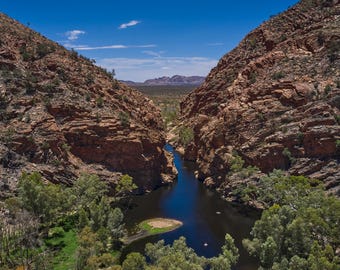 Könnte beinhalten: Ein schmaler, tiefer Fluss fließt zwischen zwei hoch aufragenden, zerklüfteten roten Felsklippen unter einem strahlend blauen Himmel. Grüne Eukalyptusbäume säumen die Ufer und eine kleine Sandinsel liegt im dunklen Wasser, mit Bergen am Horizont.
