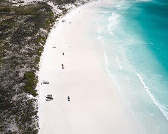 Könnte beinhalten: Eine Luftaufnahme eines langen weißen Sandstrandes, der auf türkisfarbenes Meerwasser trifft. Mehrere Fahrzeuge parken entlang der Küstenlinie, und dunkelgrüne Vegetation grenzt auf der linken Seite an den Sand.