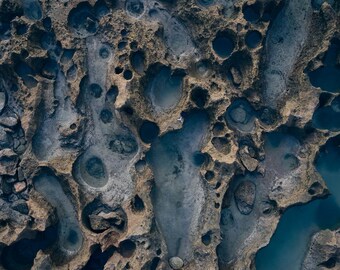 Könnte beinhalten: Eine Luftaufnahme einer rauen, porösen Felsformation mit zahlreichen natürlichen Gezeitentümpeln, die mit dunkelblauem und grauem Wasser gefüllt sind. Die texturierte, braune Steinoberfläche ist von kreisförmigen Vertiefungen durchzogen.