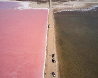 Könnte beinhalten: Eine Luftaufnahme einer langen, geraden Schotterstraße, die zwei Gewässer trennt. Links ist das Wasser in einem leuchtenden, soliden Pink gefärbt. Rechts ist das Wasser dunkel und trüb braun. Mehrere kleine Fahrzeuge parken entlang der Straße.