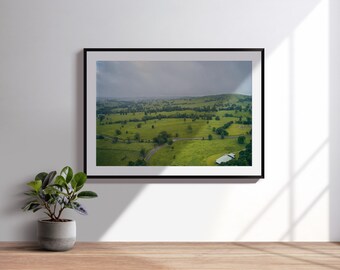 Könnte beinhalten: Ein gerahmtes Landschaftsfoto mit einer weiten Luftaufnahme von grünen Hügeln, vereinzelten Bäumen und einer kurvenreichen Straße unter einem bewölkten Himmel. Der Druck befindet sich in einem dünnen schwarzen Rahmen an einer weißen Wand.
