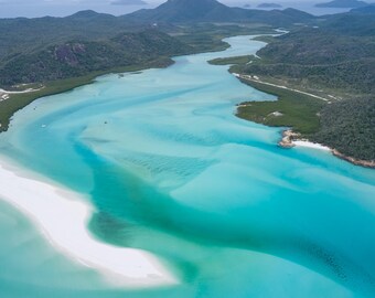 Könnte beinhalten: Eine Luftaufnahme von Hill Inlet am Whitehaven Beach mit wirbelnden Mustern aus türkisfarbenem und aquamarinblauem Wasser, das sich mit weißen Sandbänken vermischt, umgeben von dichter grüner Küstenvegetation und fernen Hügeln.