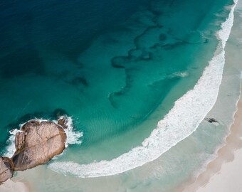 Könnte beinhalten: Eine Luftaufnahme einer Küste mit einem weißen Sandstrand, der auf türkisfarbenes und tiefblaues Meerwasser trifft. Große, glatte, hellbraune Felsen liegen im flachen Wasser, während weißer Gischt von Wellen an das Ufer spült.