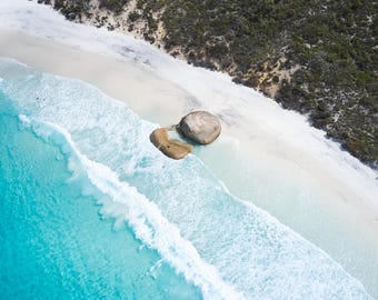 Könnte beinhalten: Eine Luftaufnahme eines weißen Sandstrandes, der auf türkisfarbene Meereswellen trifft. Zwei große, glatte, hellbraune Felsbrocken liegen am Ufer nahe der Wasserlinie, gesäumt von einer dichten Fläche aus dunkelgrüner Küstenvegetation.