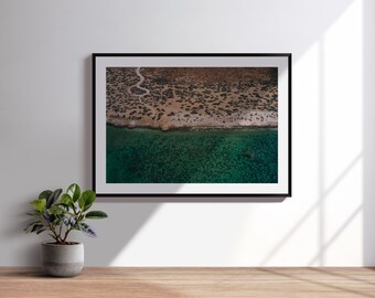Könnte beinhalten: Gerahmte Luftaufnahme einer Küstenlinie. Das Bild zeigt eine Küstenlinie mit einer Mischung aus beigem und braunem Land, das auf klares, türkisfarbenes Wasser trifft. Das Foto ist in einem schwarzen Rahmen und wird an einer weißen Wand angezeigt.